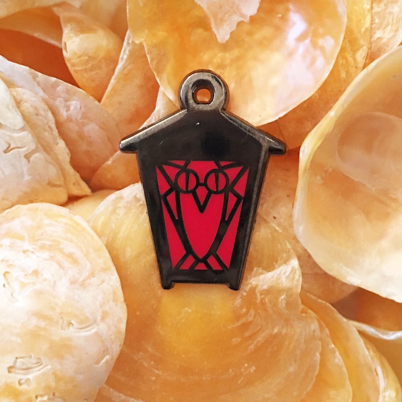 May include: A black metal lantern charm with a red owl design. The lantern has a pointed roof and a rectangular body with a window on each side. The owl has a red body and black eyes.