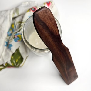 Peut inclure: Une spatule en bois brun foncé avec une extrémité arrondie, posée sur un bocal en verre rempli d'une substance blanche.