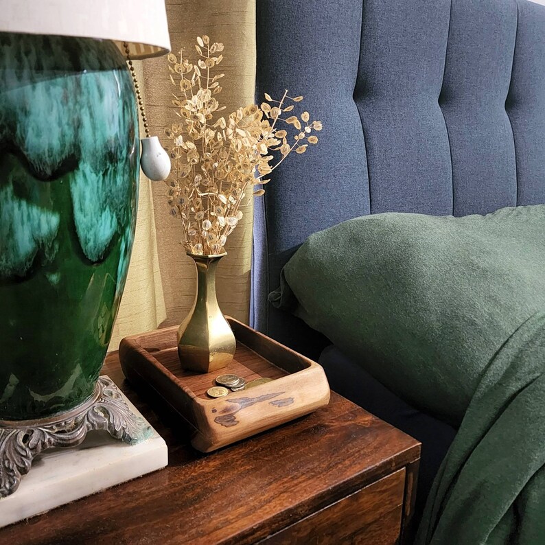 May include: A wooden tray with a decorative design sits on a dark wood table. The tray has a few coins on it. A green ceramic lamp with a white shade is in the background. A blue upholstered headboard with a green pillow is also in the background.