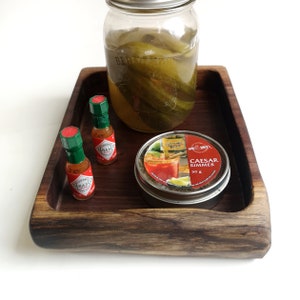 May include: A wooden tray with a glass jar of dill pickles, two small bottles of Tabasco sauce, and a small tin of Caesar Rimmer.
