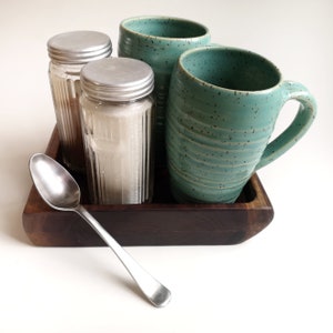 May include: A wooden tray with two teal mugs and two glass jars with silver lids. A silver spoon is on the tray.