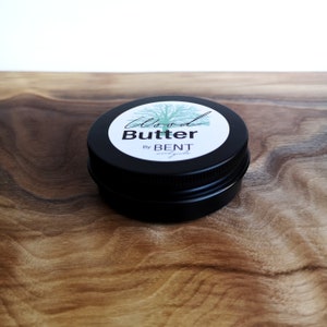 May include: A small black tin container with a white label that reads "Wood Butter by BENT" and a green tree illustration.