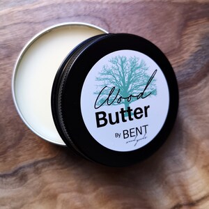 May include: A black tin with a white label that says "Wood Butter by BENT wood goods". The label has a green tree illustration. The tin is open and the inside is filled with a white substance.