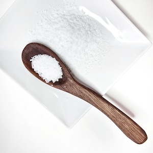 May include: A wooden spoon filled with white salt on a white square plate. The spoon is made of dark wood and has a curved handle.