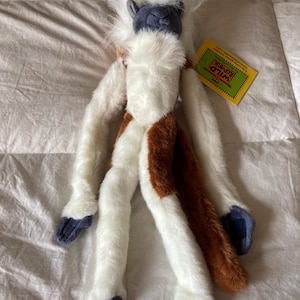 May include: A plush lemur toy with long white fur, a dark gray face, and dark gray hands and feet. The stuffed animal features patches of brown fur on its body and a long brown tail. A yellow Wild Republic brand tag is attached to the ear.