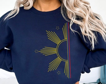 May include: A navy blue crewneck sweatshirt featuring a graphic of a yellow sunburst design on the left side. The sun graphic includes rays and three small stars. A vertical stripe in red, yellow, and blue runs along the right side of the sun.