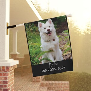 May include: A rectangular garden flag featuring a photograph of a white dog sitting in green foliage. Below the image, a black horizontal band displays the name Bella in a white script font, followed by the text RIP 2005-2024 in a sans-serif font.