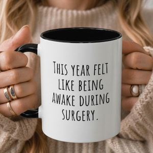 May include: White ceramic mug with a black interior and handle. The mug features the text "THIS YEAR FELT LIKE BEING AWAKE DURING SURGERY" in black, sans-serif font. The mug is held by a person wearing a beige sweater.
