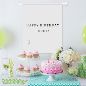 May include: A white fabric wall hanging banner with the black text HAPPY BIRTHDAY SOPHIA, displayed above a table featuring a two-tier cupcake stand, a pink frosted cake with two candles, green polka dot napkins, and a bouquet of white flowers.