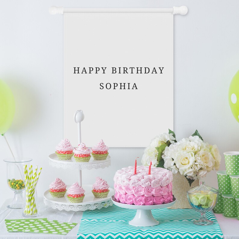 May include: A white fabric wall hanging banner with the black text HAPPY BIRTHDAY SOPHIA, displayed above a table featuring a two-tier cupcake stand, a pink frosted cake with two candles, green polka dot napkins, and a bouquet of white flowers.