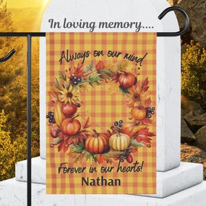 May include: A garden flag with a fall theme. The flag features a wreath of pumpkins, sunflowers, and leaves on a yellow and orange plaid background. Text reads "Always on our mind, Forever in our hearts!" and the name "Nathan".
