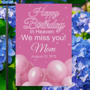 May include: A pink garden flag with the text "Happy Birthday In Heaven, We miss you! Mom, August 01, 1973." The flag features white script and pink balloons at the bottom. Blue flowers are in the background.