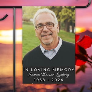 May include: A rectangular garden flag featuring a portrait of a smiling person with gray hair and glasses, wearing a black sweater over a light patterned shirt. Below the portrait, black text reads IN LOVING MEMORY James Thomas Ludwig 1958 - 2024.