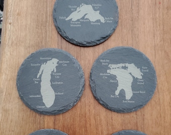 May include: Five round, dark gray slate coasters with etched maps of the Great Lakes region. Each coaster features a different lake with city names. The coasters are arranged on a wooden surface.