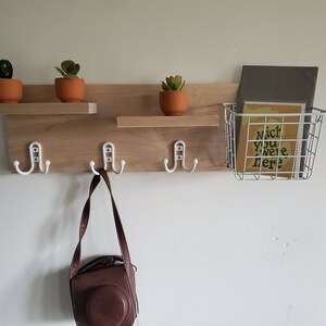May include: A wooden wall shelf with three hooks and a wire basket. The shelf has three potted succulents and a brown leather bag hanging from one of the hooks. The basket contains a book with the text "Nice you were here".