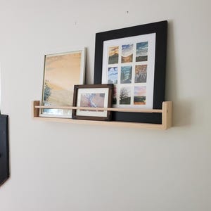 May include: A wooden shelf with three framed pictures on it. The shelf is mounted on a white wall. The pictures are all different sizes and have different images in them.