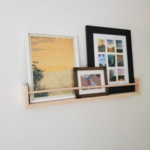 May include: A light wood shelf with three framed pictures. The shelf is mounted on a white wall. The pictures are all different sizes and have different subjects. The largest picture is a landscape scene with a yellow sky and a blue body of water. The middle picture is a landscape scene with a river and trees. The smallest picture is a landscape scene with a mountain range.