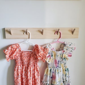 May include: Two floral print dresses hanging on a wooden coat rack with hooks. The dress on the left is orange with white flowers and the dress on the right is white with pink, yellow, and purple flowers.