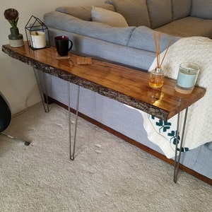 May include: A long, narrow console table featuring a natural live-edge wood slab top with a polished finish, supported by four slender, silver-toned metal hairpin legs. The table surface displays a rich brown wood grain and textured bark edges.