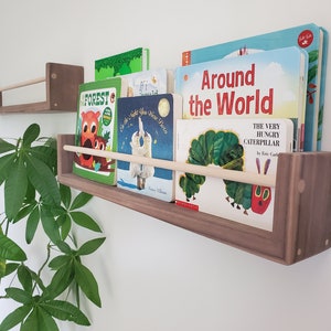 May include: Two wooden shelves with a dowel rod for holding books. The shelves are mounted on a white wall. The top shelf is empty. The bottom shelf is filled with children's books, including "The Very Hungry Caterpillar" and "Around the World".