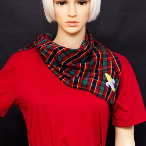 May include: A red t-shirt and light blue denim jeans are worn by a mannequin. A plaid scarf in red, green, yellow, and navy blue is draped around the mannequin's neck. A small, embroidered banana pin is attached to the scarf.