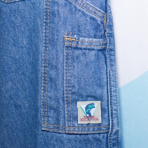 Puede incluir: Primer plano de un pantal&oacute;n de trabajo de mezclilla azul con m&uacute;ltiples bolsillos y costuras visibles. Un peque&ntilde;o parche rectangular en el bolsillo presenta un gr&aacute;fico de un pez y el texto "WEAR KICKWEAR".