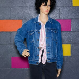 May include: A classic blue denim jacket with button closures and chest pockets. The jacket is worn over a red and white striped shirt and dark jeans. The mannequin is positioned in front of a gray brick wall with colorful painted squares.