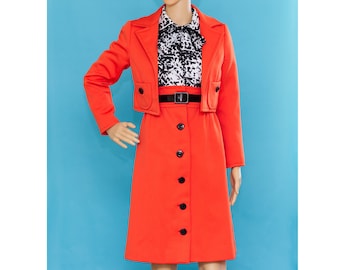 May include: A vintage orange skirt suit on a mannequin. The suit includes a cropped jacket with black buttons and a matching skirt. A black and white patterned blouse and a black belt complete the outfit. The mannequin has a blonde wig and is set against a blue background.