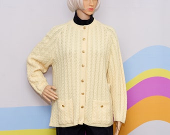 May include: A light yellow cable-knit cardigan with gold-colored buttons and two front pockets. The long-sleeved sweater has a round neckline and a classic design. The cardigan is displayed on a mannequin.