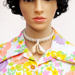 May include: A mannequin wearing a white shirt with a vibrant floral pattern in orange, yellow, and purple. The shirt features a collar and buttons. A white beaded necklace with shell pendants is also visible.