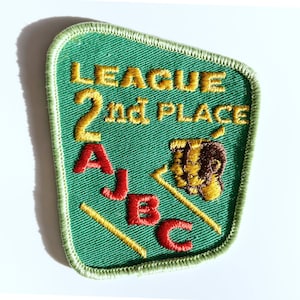 May include: A green embroidered patch with the words "LEAGUE 2nd PLACE" in gold thread. The patch also features red letters "A, J, B, C" and a gold and brown image of a bear. The patch has a green border.