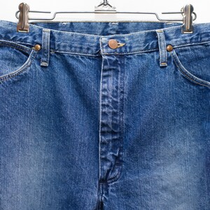 May include: Close-up of blue denim jeans hanging on a metal hanger. The jeans feature a button and zip closure, front pockets, and copper-coloured rivets. The denim has a faded appearance.