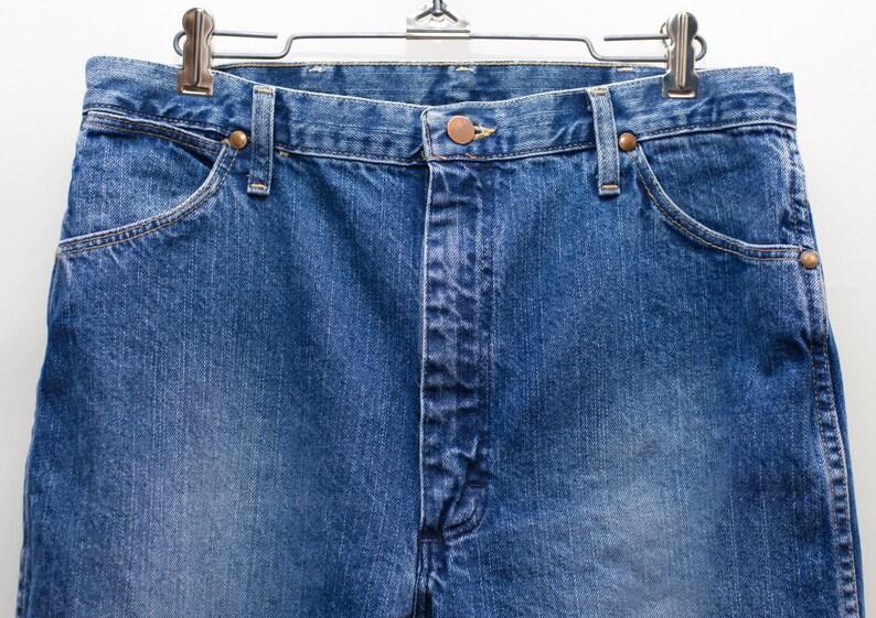May include: Close-up of blue denim jeans hanging on a metal hanger. The jeans feature a button and zip closure, front pockets, and copper-coloured rivets. The denim has a faded appearance.