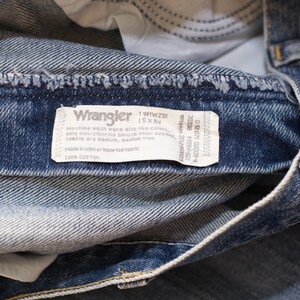 May include: Close-up of blue denim Wrangler jeans. The jeans have a faded wash and a white tag with the Wrangler logo and care instructions. The tag also includes the size 15 x 34.