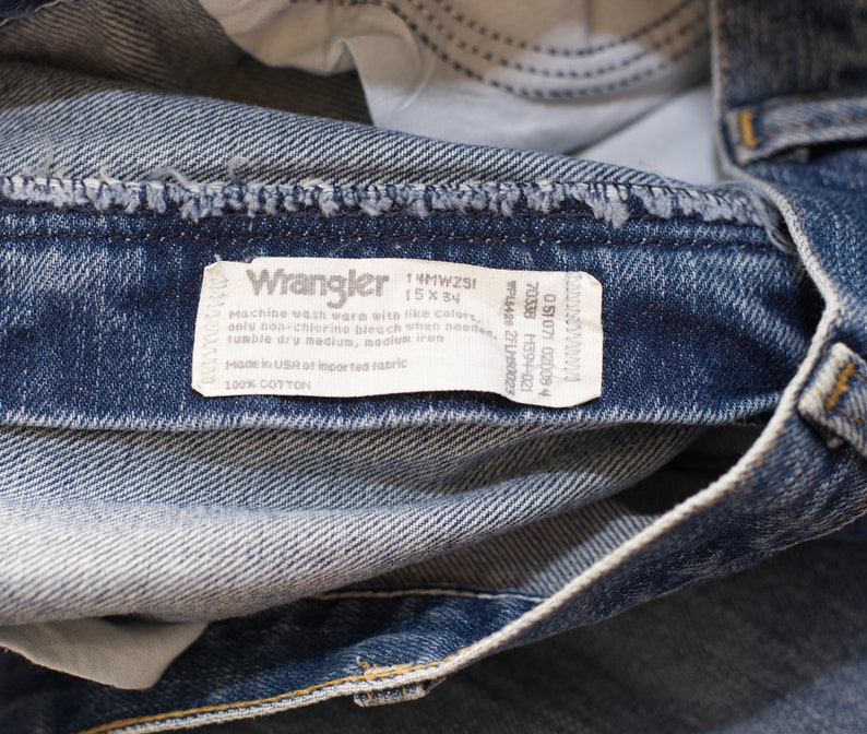 May include: Close-up of blue denim Wrangler jeans. The jeans have a faded wash and a white tag with the Wrangler logo and care instructions. The tag also includes the size 15 x 34.