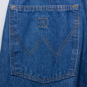 Puede incluir: Primer plano de un bolsillo de vaquero azul. El bolsillo presenta un dise&ntilde;o de costura "W" distintivo en hilo marr&oacute;n, con costuras a juego en los bordes. Un peque&ntilde;o detalle cuadrado cosido est&aacute; encima del dise&ntilde;o "W".