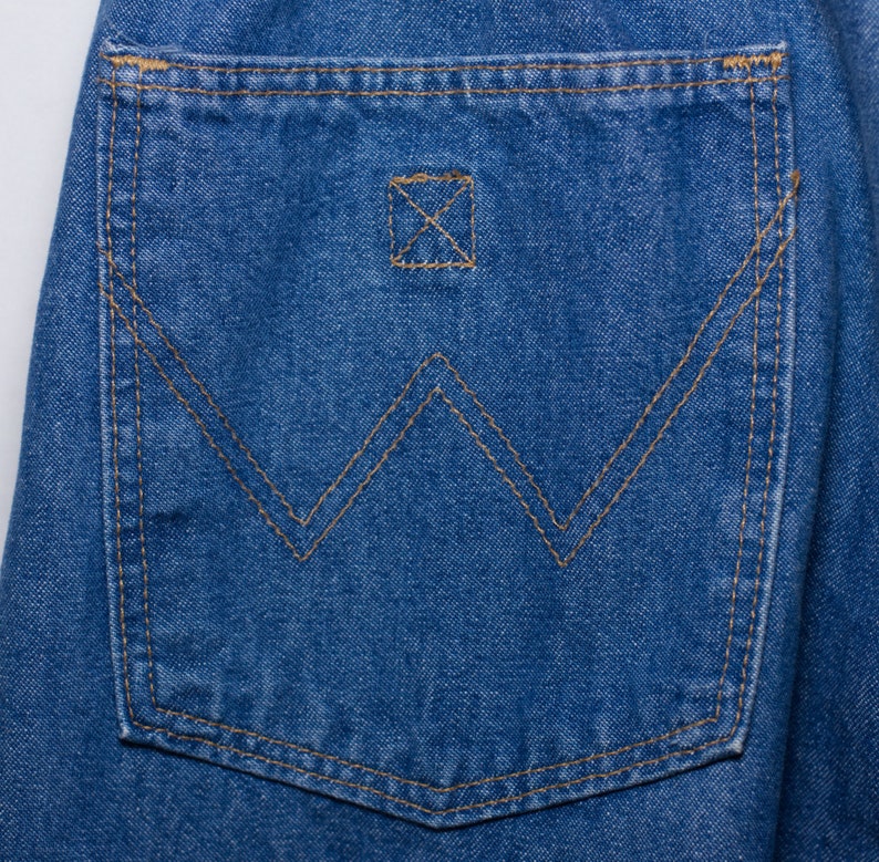 Puede incluir: Primer plano de un bolsillo de vaquero azul. El bolsillo presenta un dise&ntilde;o de costura "W" distintivo en hilo marr&oacute;n, con costuras a juego en los bordes. Un peque&ntilde;o detalle cuadrado cosido est&aacute; encima del dise&ntilde;o "W".
