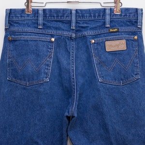 May include: Close-up of the back of blue denim jeans with the Wrangler logo on a brown leather patch. The jeans have two back pockets with a signature W-shaped stitching design and brass rivets. The jeans are hanging on a metal hanger.