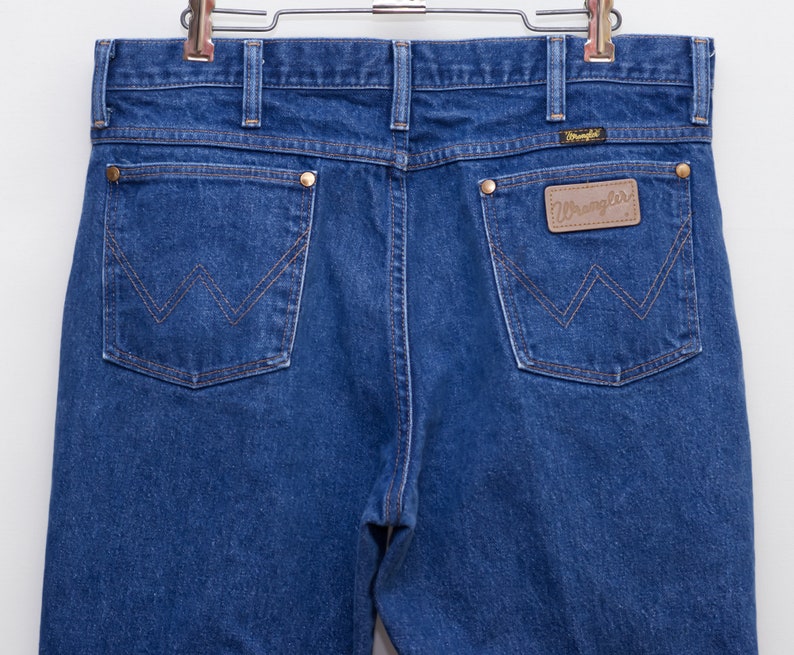 May include: Close-up of the back of blue denim jeans with the Wrangler logo on a brown leather patch. The jeans have two back pockets with a signature W-shaped stitching design and brass rivets. The jeans are hanging on a metal hanger.