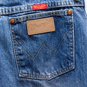 May include: Close-up of blue denim jeans showing a back pocket with the Wrangler logo on a brown leather patch. The pocket has a classic W-shaped stitch design. A red Wrangler tag is visible.