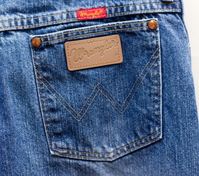 May include: Close-up of blue denim jeans showing a back pocket with the Wrangler logo on a brown leather patch. The pocket has a classic W-shaped stitch design. A red Wrangler tag is visible.