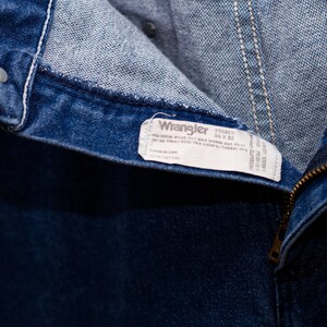 May include: Close-up of blue denim jeans with a zipper and a button closure. The jeans have a white tag that reads "Wrangler" with the size 36 x 32. The jeans are made of cotton.