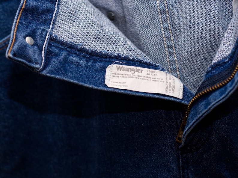 May include: Close-up of blue denim jeans with a zipper and a button closure. The jeans have a white tag that reads "Wrangler" with the size 36 x 32. The jeans are made of cotton.