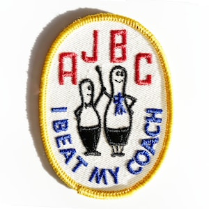 May include: Oval embroidered patch with a white background, yellow border, and the text "A J B C I BEAT MY COACH" in red and blue. Two cartoon figures are depicted, one with a blue bow tie.