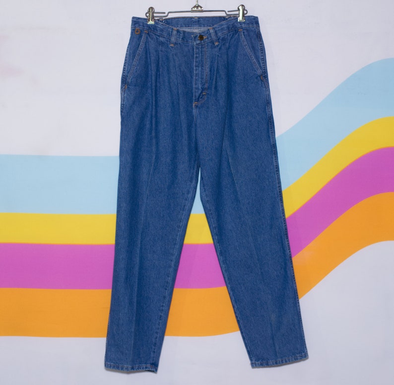 Puede incluir: Pantalones vaqueros azules con la parte delantera plisada, colgados de una percha met&aacute;lica. Los pantalones tienen un corte cl&aacute;sico de pierna recta y cuentan con bolsillos y cierre de bot&oacute;n. El fondo tiene un dise&ntilde;o de rayas de arco&iacute;ris colorido.