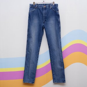 May include: Blue denim jeans hanging on a metal hanger. The jeans feature a classic five-pocket design, a button and zip closure, and a straight leg. The denim has a faded wash, giving it a worn appearance.