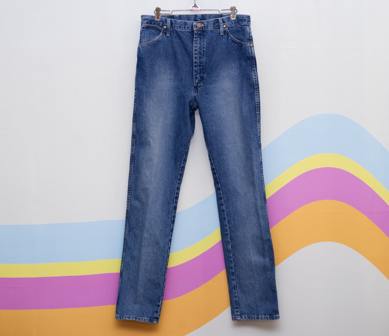 May include: Blue denim jeans hanging on a metal hanger. The jeans feature a classic five-pocket design, a button and zip closure, and a straight leg. The denim has a faded wash, giving it a worn appearance.