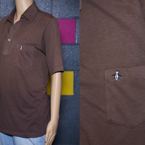 May include: Brown polo shirt with a pocket on the chest. The shirt has short sleeves and a collar. A small embroidered penguin logo is on the pocket. The shirt is displayed on a mannequin.