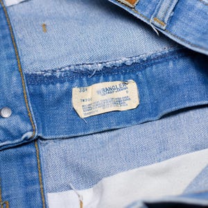 Puede incluir: Primer plano de una prenda de mezclilla azul claro, posiblemente jeans o una chaqueta, con una etiqueta visible. La etiqueta dice "WRANGLER NO-FAULT DENIMS" e incluye informaci&oacute;n de talla, "38". La prenda tiene una textura y costuras de mezclilla cl&aacute;sicas.