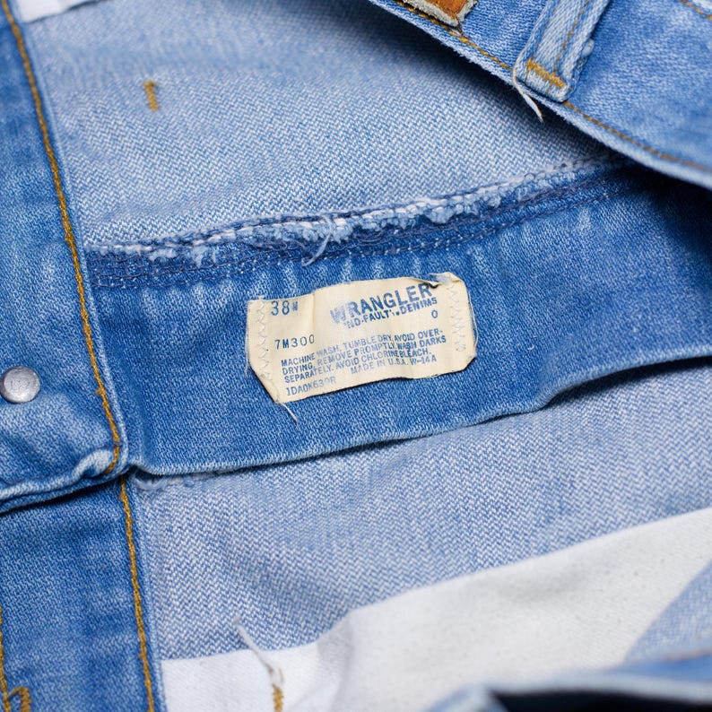 Puede incluir: Primer plano de una prenda de mezclilla azul claro, posiblemente jeans o una chaqueta, con una etiqueta visible. La etiqueta dice "WRANGLER NO-FAULT DENIMS" e incluye informaci&oacute;n de talla, "38". La prenda tiene una textura y costuras de mezclilla cl&aacute;sicas.